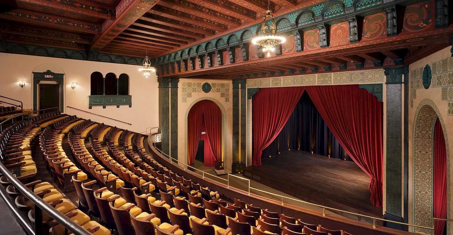 A grand vintage theater with wooden ceiling beams, red velvet curtains, ornate decorations, and many wooden seats facing a small stage. End.