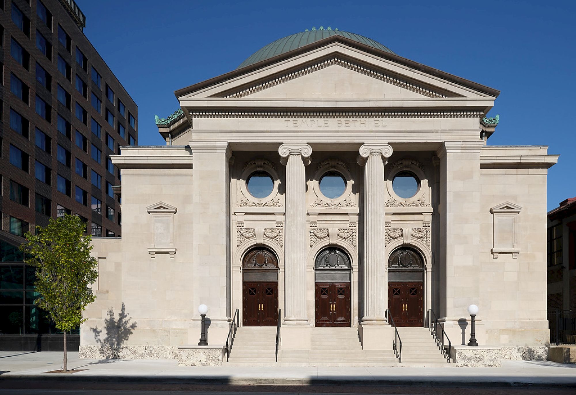 A neoclassical building with three arched wooden doors, four columns, and a domed roof, set under a clear blue sky.