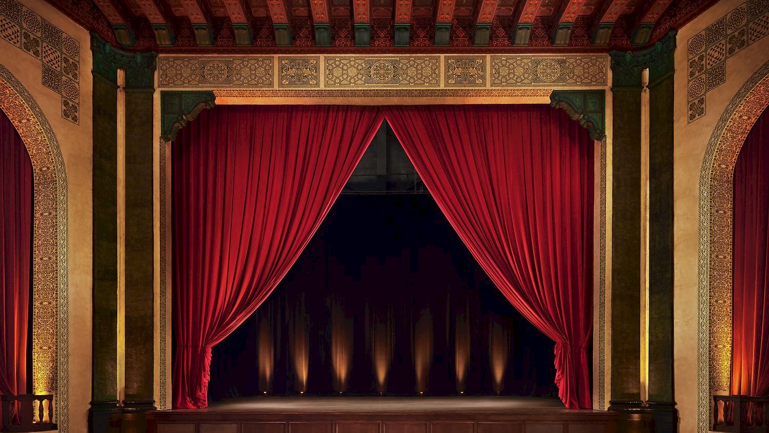 A grand theater stage with red curtains, ornate gold and green decor, and soft backlighting in a historic, elegant interior.