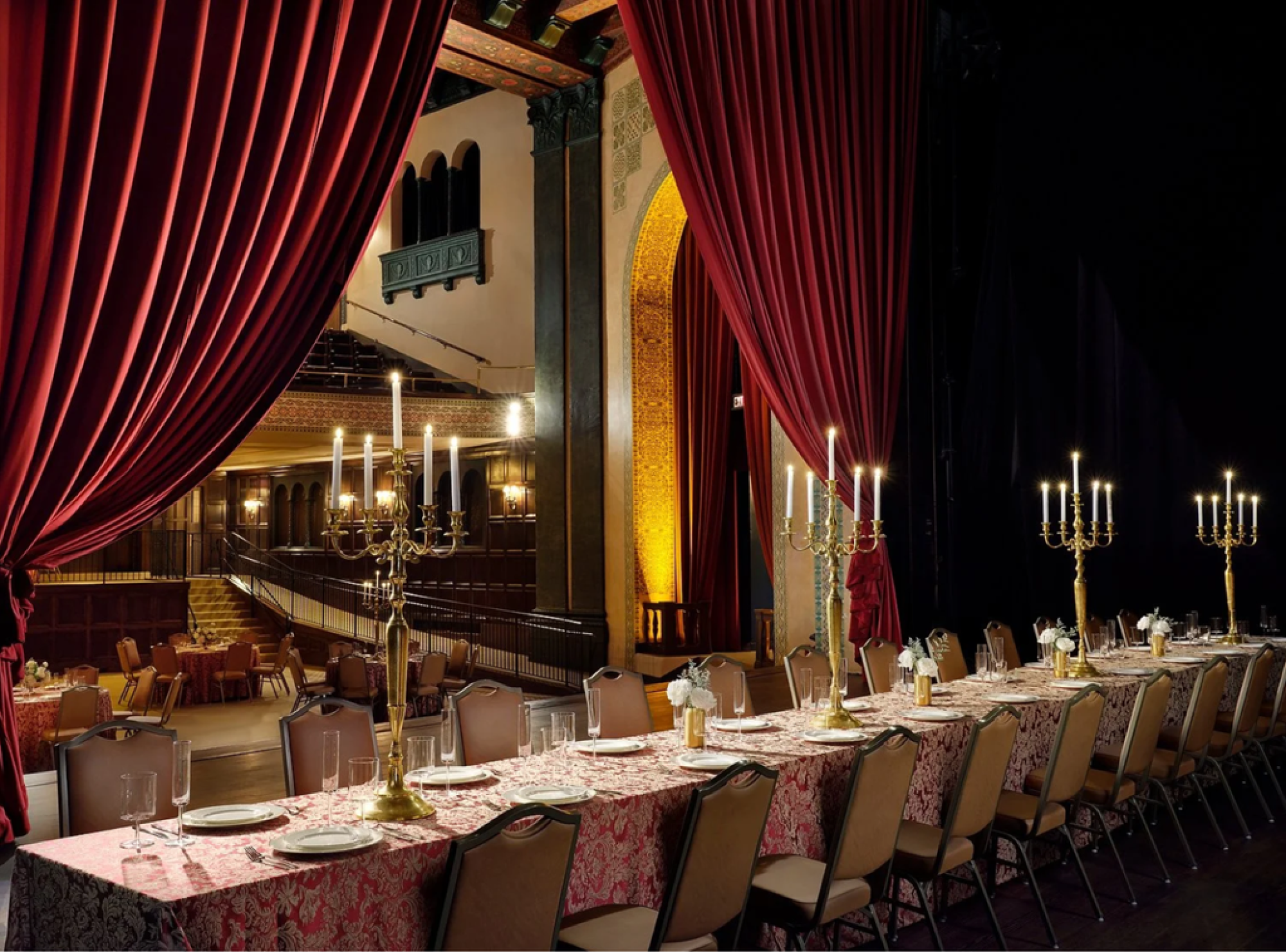 A long table set for an elegant event is framed by red curtains and candelabras, with ornate architecture in the background.