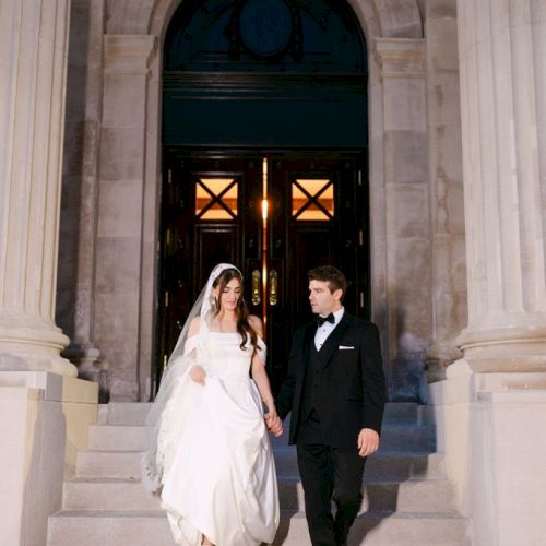 A bride and groom dressed in wedding attire descend stone stairs in front of an elegant building entrance.