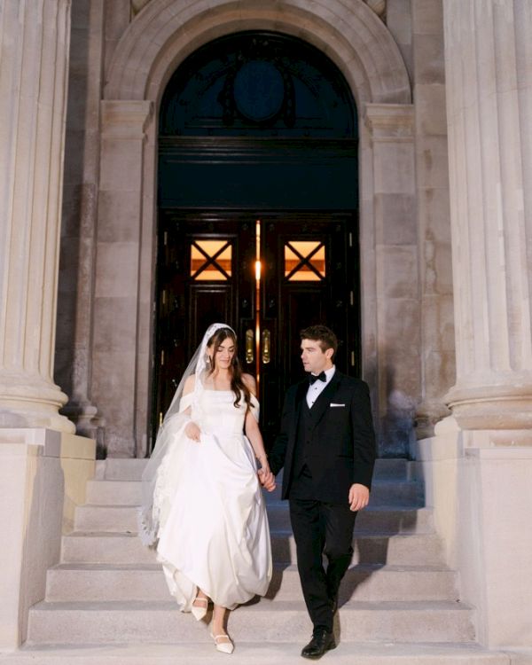 A bride and groom dressed in wedding attire descend stone stairs in front of an elegant building entrance.