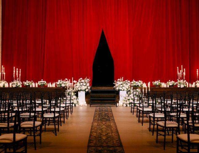 A wedding ceremony setup with rows of chairs, decorative candles, and flowers, in front of a red curtain on a carpeted aisle.