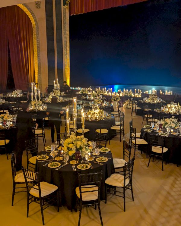 The image shows an elegantly set up banquet hall with round tables, black chairs, candles, and floral arrangements, ready for an event.