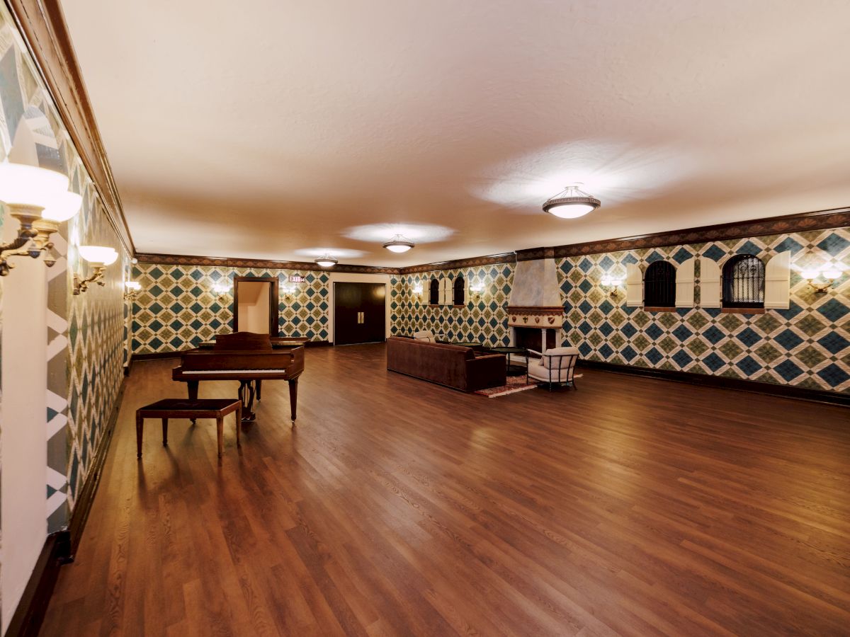 The image shows a spacious room with wooden flooring, patterned walls, a piano, and seating area, under soft lighting fixtures.