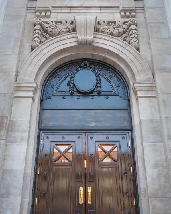 A grand wooden door with intricate carvings and gold handles, set in an ornate stone archway with decorative elements above.