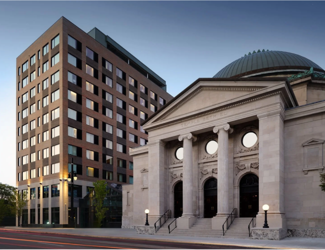 The image shows a modern building next to a classical structure with columns and a dome, possibly a historical or cultural site in an urban setting.