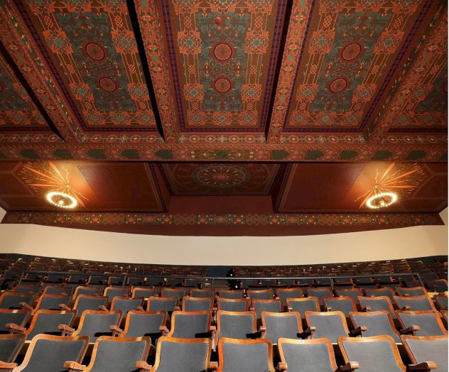 The image shows a theater or lecture hall with tiered seating and an ornate, decorative ceiling featuring detailed patterns and warm colors.