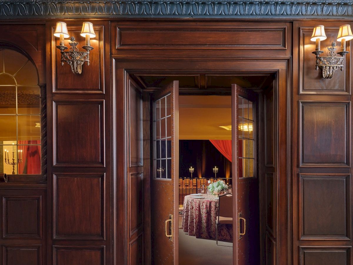 A grand wooden doorway opens to a warmly lit room with a plush, patterned table setting seen through glass doors.