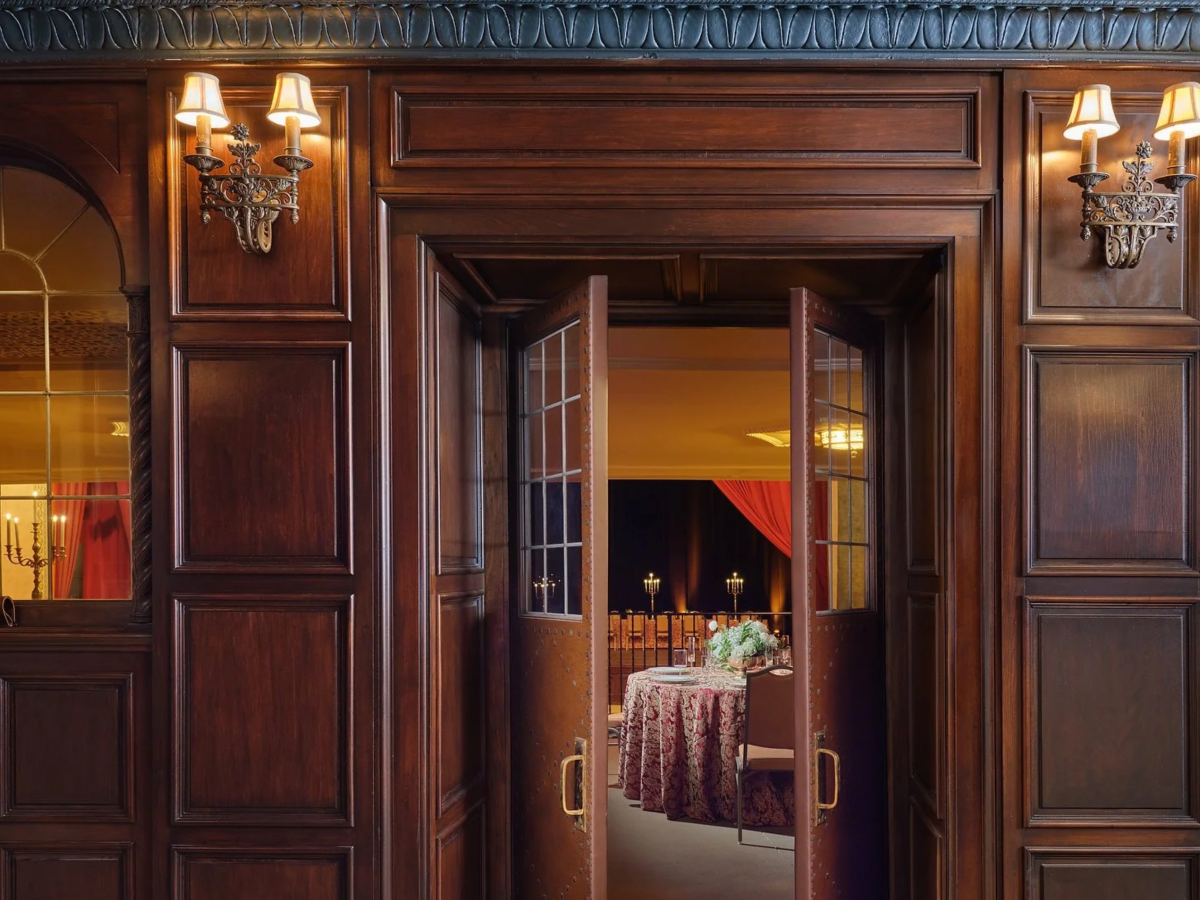 A wooden doorway with glass-paneled doors opens to a warmly lit room adorned with a table and chairs, beneath stylish wall sconces.